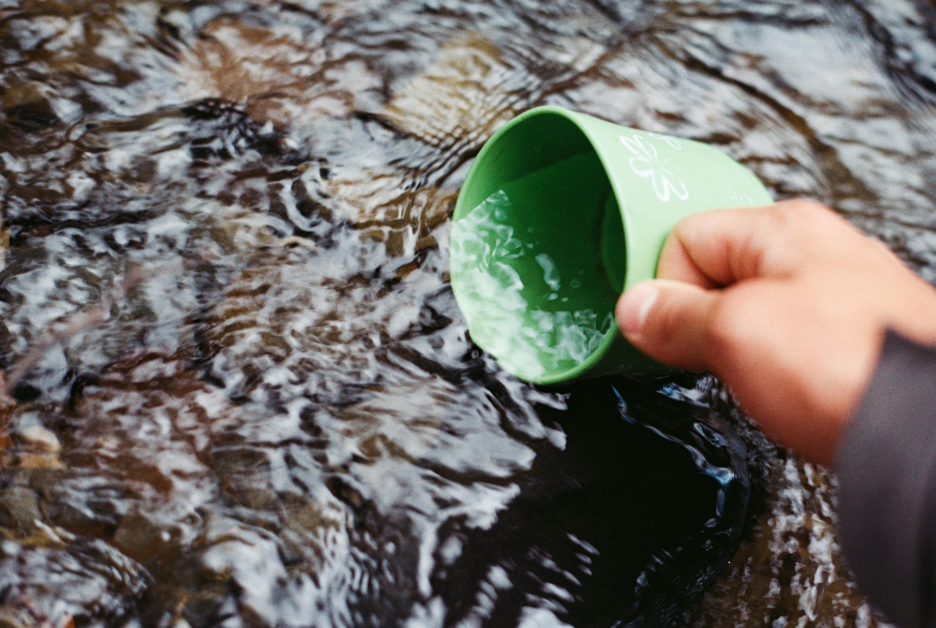 A person scooping water from a stream with a green cup